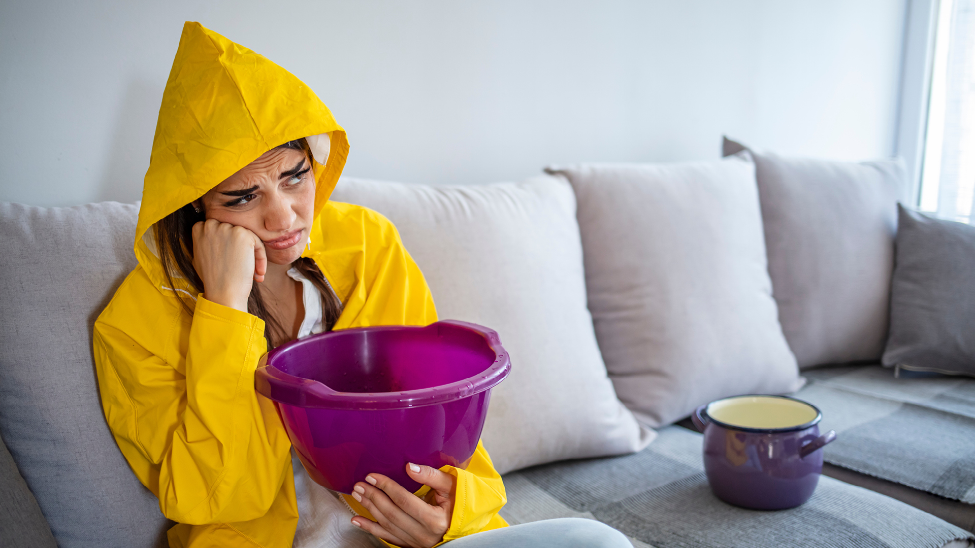 Femme en manteau assise sur son canapé regardant la fuite de sa toiture et tenant un seau dans ses bras