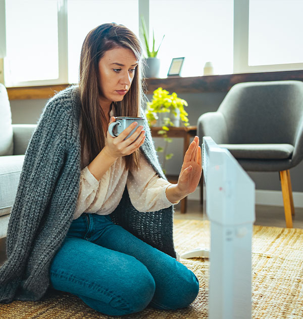 Femme à genoux ayant froid et essayant de se réchauffer avec un plaid, tasse chaude et radiateur